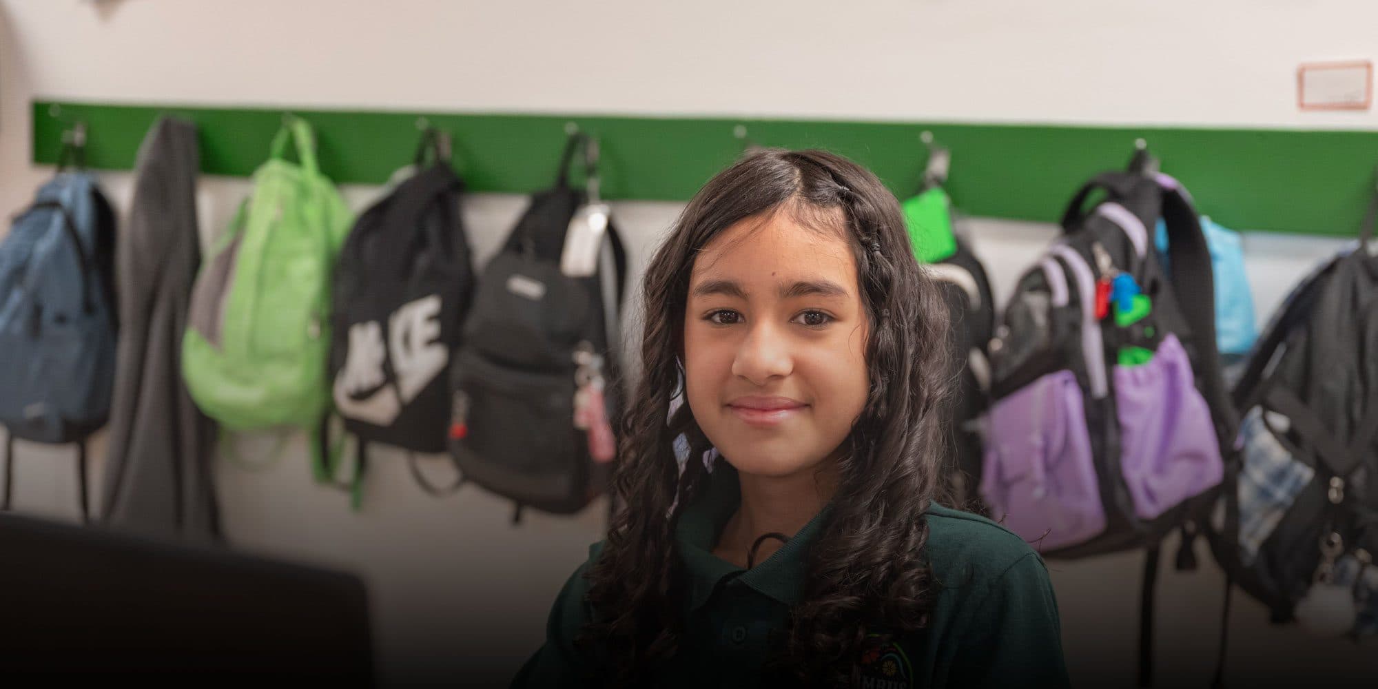 Smiling student in a green school polo in front of backpacks on classroom hooks.